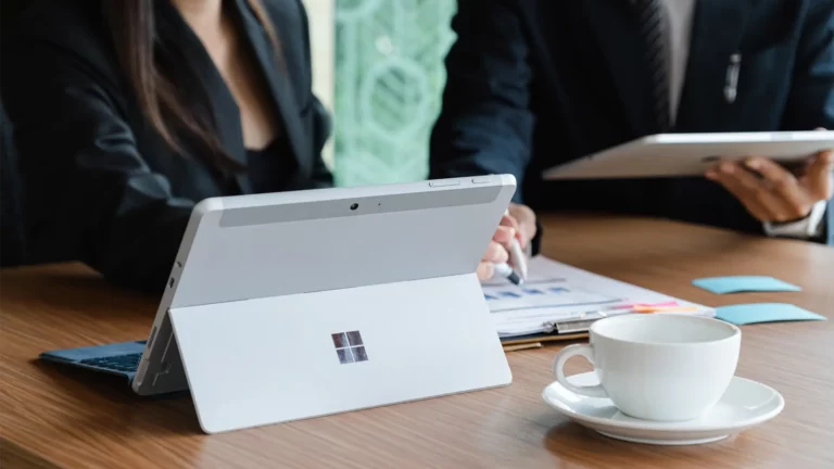 A Microsoft tablet rests on a wooden table beside a coffee cup as professionals discuss documents highlighting Microsoft 365s collaborative benefits