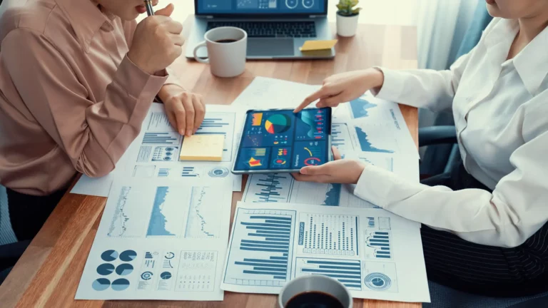 Two professionals analyzing data on a tablet surrounded by printed graphs and reports focusing on cost savings in outsourcing