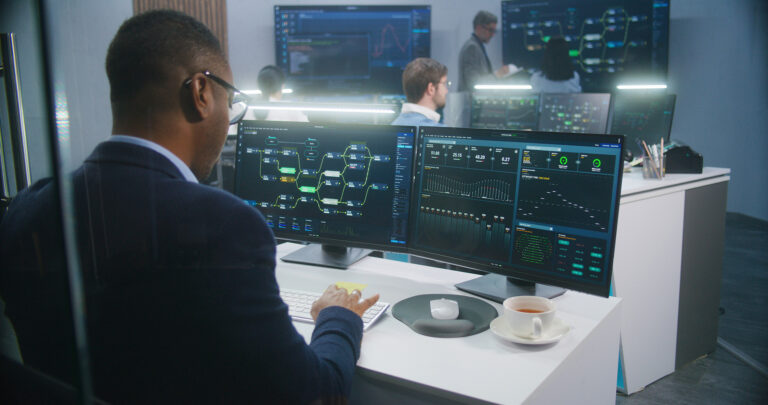 Big data scientist analyzes server data on multiple screens in a modern monitoring room with colleagues discussing in the background