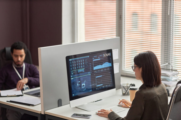Two office workers at desks with a partition focusing on data analysis displayed on their computer screens