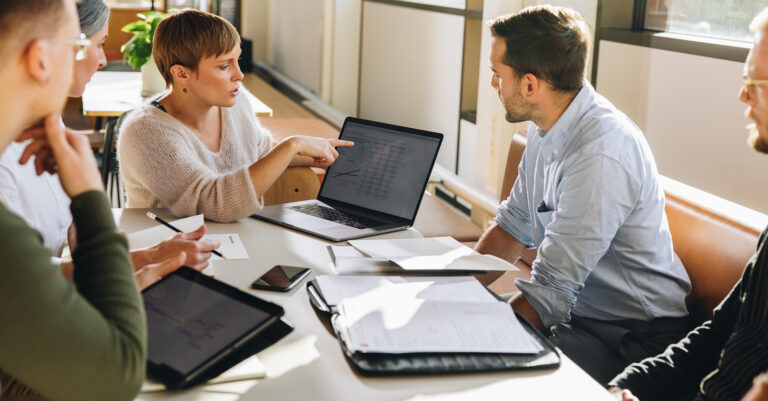 Female executive discussing accounts payable data with team during business meeting pointing at laptop screen