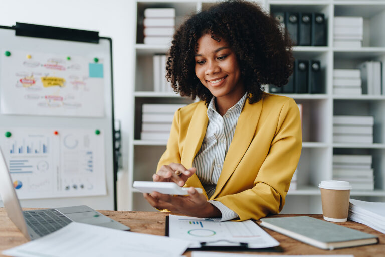 Young woman in a yellow blazer calculates expenses with a calculator surrounded by financial documents and a laptop focused on budgeting