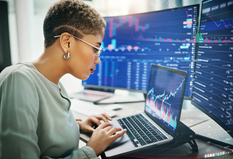 Businesswoman analyzing investment graphs on a laptop surrounded by multiple screens displaying financial data and analytics