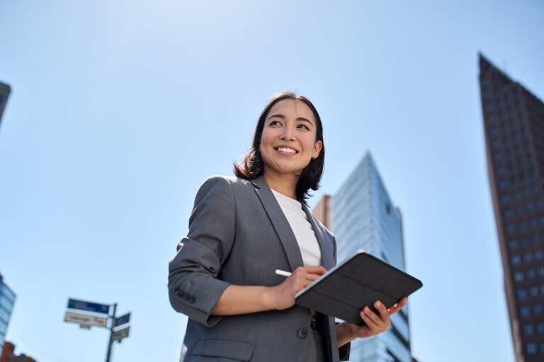 Smiling young Asian businesswoman in a gray suit holding a tablet standing in an urban setting under a clear blue sky