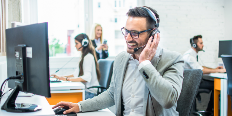 Customer service representative wearing a headset smiling while assisting clients at a busy help desk office environment