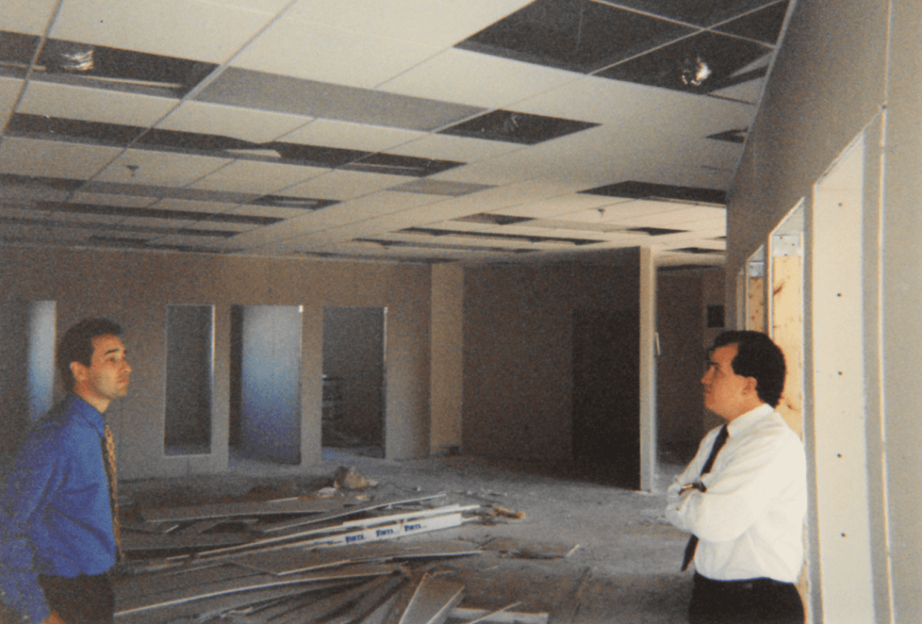 Two men in an unfinished office space discussing plans amidst construction debris and exposed ceiling tiles