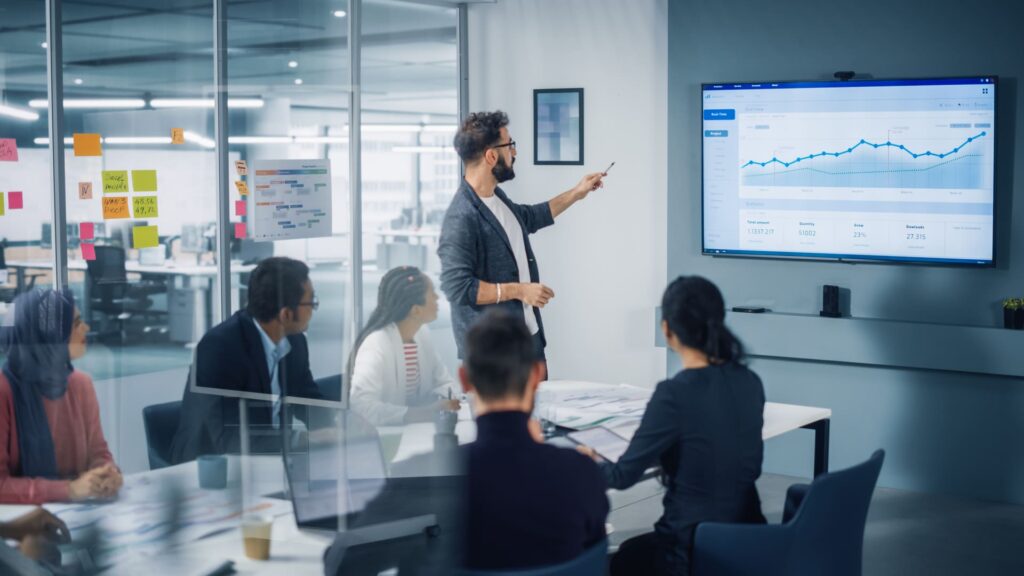 A team in a modern office discusses finance transformation with a presenter pointing at a data driven dashboard on a screen