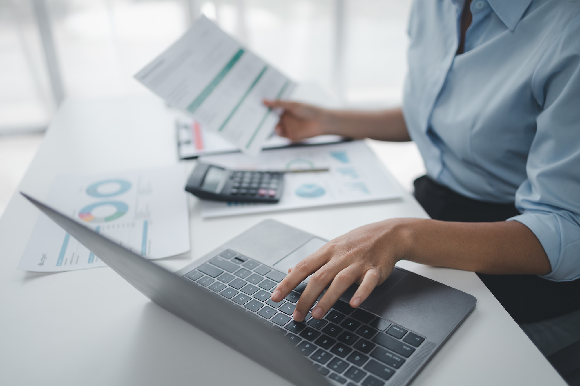 Accountant reviewing financial statements and budget documents while working on a laptop at a desk focused on auditing tasks