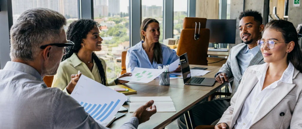 A diverse team discusses finance benchmarking metrics during a meeting analyzing charts and reports in a modern office