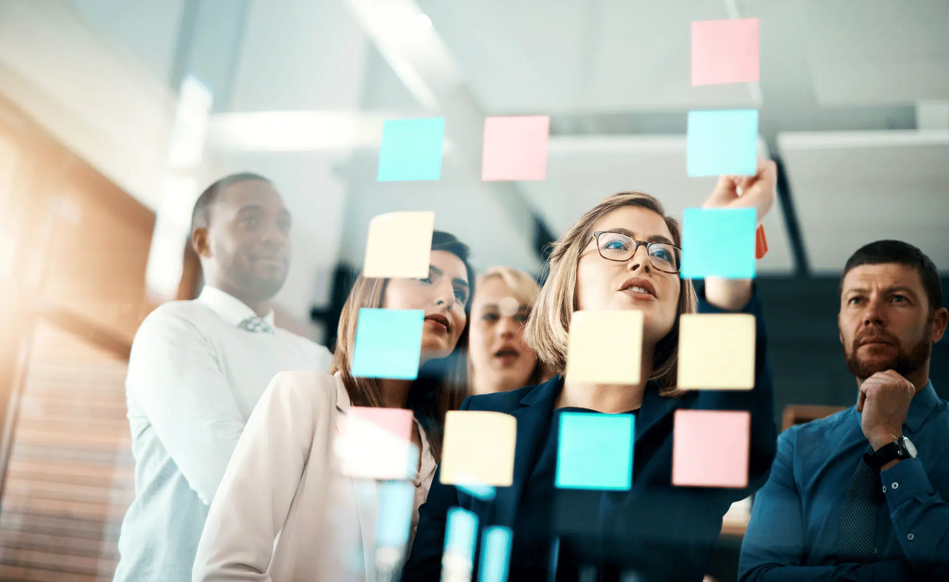 Group of diverse professionals in a modern office brainstorming and collaborating using colorful sticky notes on a glass wall The scene conveys agile teamwork strategic planning and innovation in a business environment