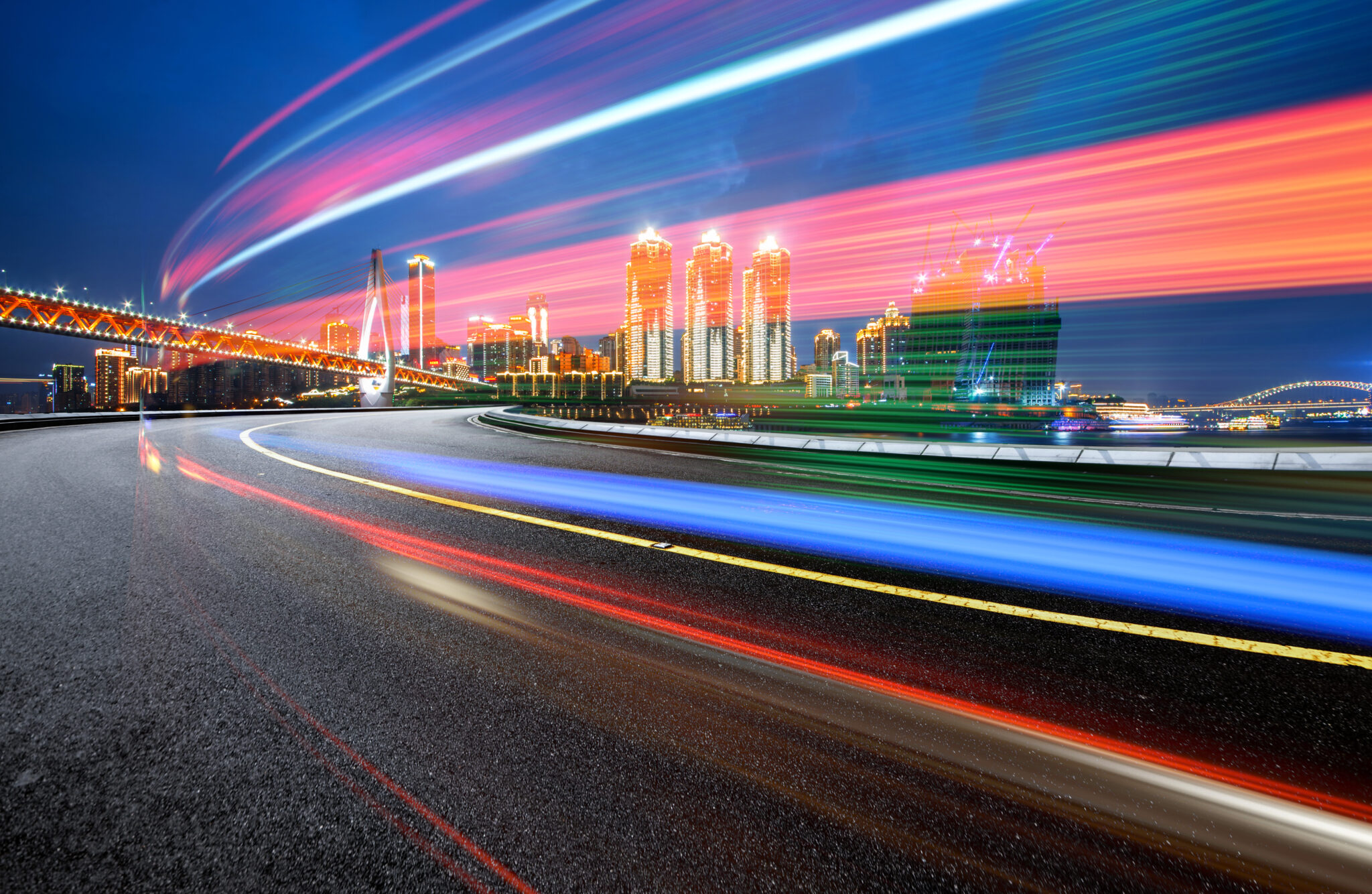 Nighttime cityscape with vibrant light trails from vehicles on a curved road passing under a lit up bridge with towering skyscrapers in the background