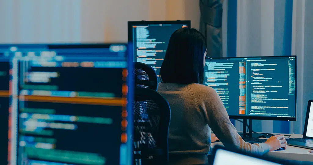 Female cybersecurity analyst working with code on multiple monitors in a dark office