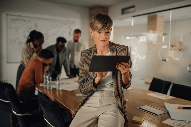 HR leader using tablet in modern office with team collaborating in the background discussing AI in human resources strategies