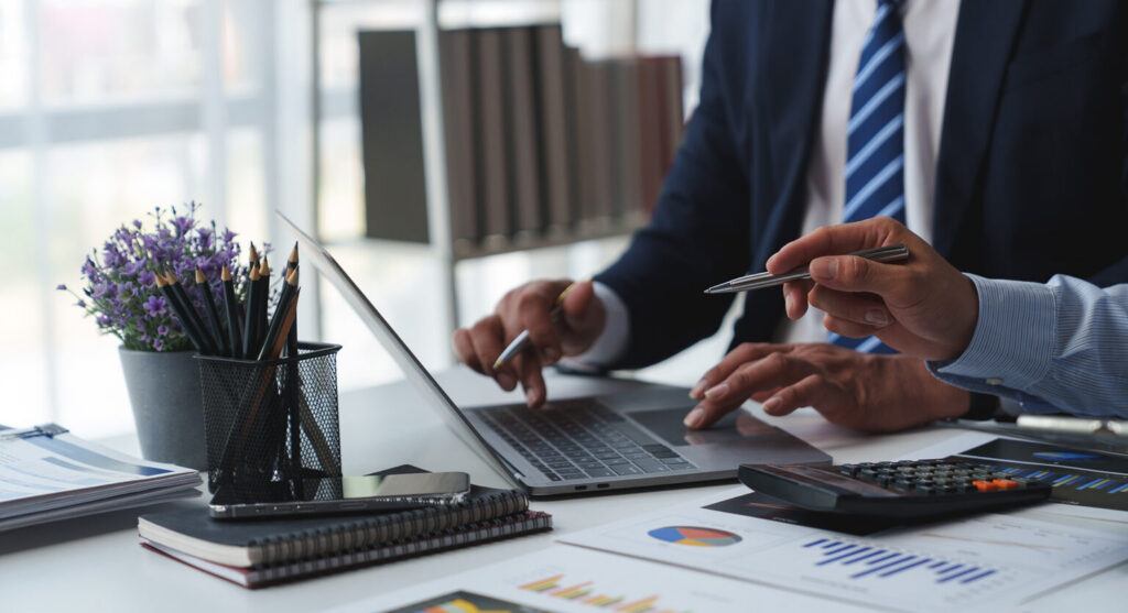 Two finance professionals in business attire collaborating over financial reports and a laptop using a calculator and pens to analyze data The scene represents accounts payable best practices such as rigorous invoice auditing data accuracy and strategic financial oversight