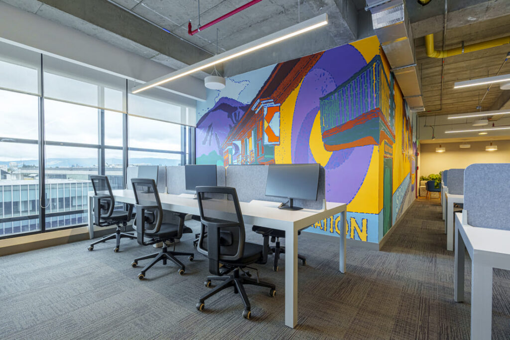 Solinoff - Auxis-14 1 1 - Auxis Bright and modern office space featuring workstations and a colorful mural symbolizing Auxis' growth in Bogotá.