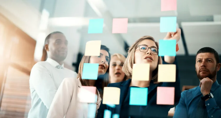 Group of diverse professionals in a modern office brainstorming and collaborating using colorful sticky notes on a glass wall. The scene conveys agile teamwork, strategic planning, and innovation in a business environment.