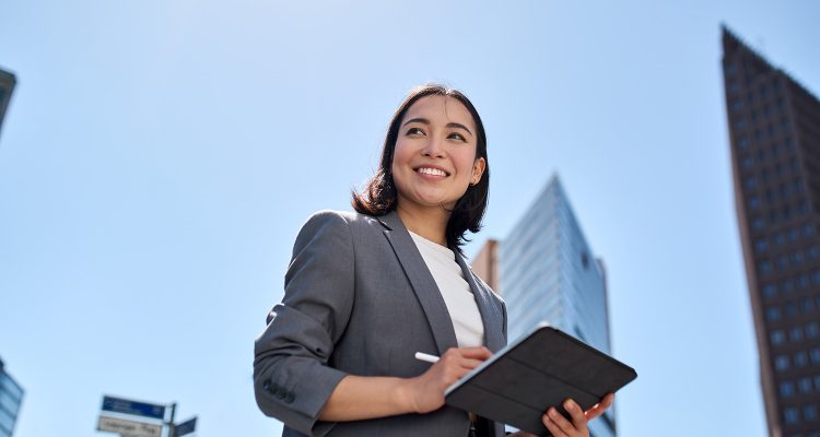 Smiling young Asian business woman leader entrepreneur, professional manager holding digital tablet computer using software applications standing on the street in big city on sky background.