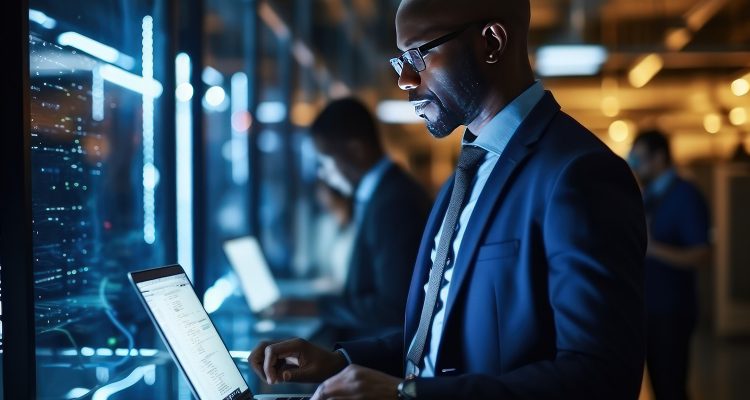 Serious African American man with laptop stands in the server room. Collection and storage of large amounts of data. Checks the operation of servers and automation.