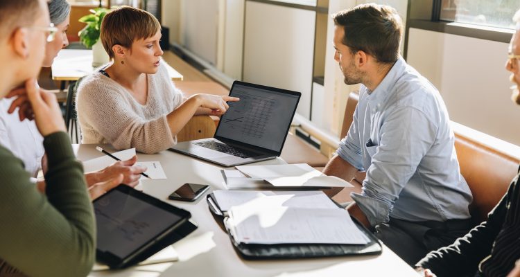 Manager pointing at laptop screen and talking to colleagues during business meeting. Female executive showing data to team in meeting.