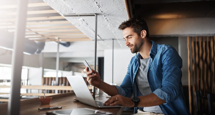 Man in coffee shop, laptop and smartphone, small business and entrepreneur in hospitality industry and connect. Male owner, cafe franchise and check email or social media with digital admin on pc.